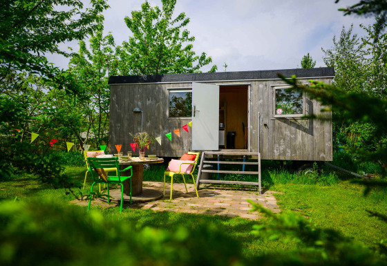 Charmante tiny house Pipowagen avec porte ouverte, chaises colorées et fanions dans un jardin verdoyant.