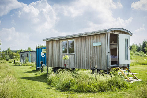 Foto des Tiny House Pipowagen aus Holz, mit offener Tür, auf einer grünen Wiese und sonnigem Himmel.