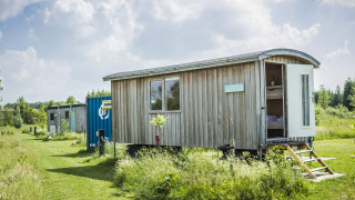 Foto della tiny house Pipowagen in legno su ruote, porta aperta, in un prato verde sotto il cielo soleggiato.