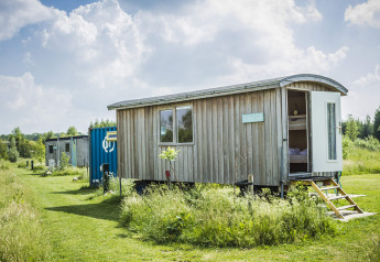 Foto della tiny house Pipowagen in legno su ruote, porta aperta, in un prato verde sotto il cielo soleggiato.