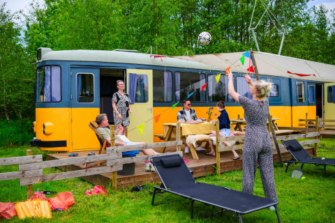 Gezin zit buiten bij tiny house Streetcar met tuinmeubilair en kleurrijke slingers in het groen.