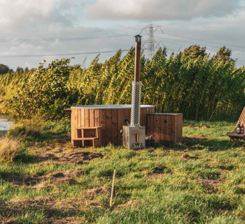Bañera de madera al aire libre junto a una tiny house, en césped verde y rodeada de vegetación alta.