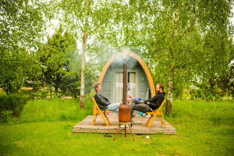 To personer sidder foran en træhytte ved Log Cabin pod, omgivet af grønne træer og natur.