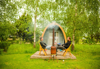 Two people relax outside a Log Cabin pod, sitting on chairs surrounded by trees and lush green grass.