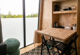 Interior de tiny house con mesa de madera, sillas y ventanales en Birdhouse, Netl Camping Kallumaan, Países Bajos.