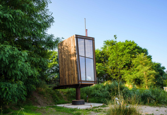 Una tiny house chiamata Birdhouse, rialzata su una colonna, circondata da vegetazione verde e alberi.