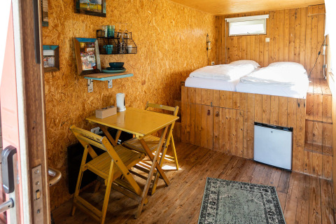 Vista interior de una pequeña casa contenedor con suelos de madera, mesa de comedor y cama en Netl Camping Kallumaan.
