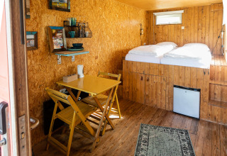 Vista interior de una pequeña casa contenedor con suelos de madera, mesa de comedor y cama en Netl Camping Kallumaan.