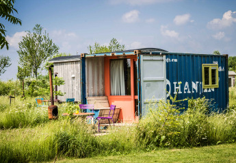 Et farverigt tiny house bygget af en shipping container, omgivet af grønt græs og vilde blomster.