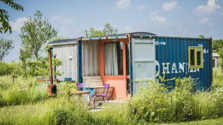 A colorful tiny house made from a shipping container sits in a grassy field surrounded by wildflowers.