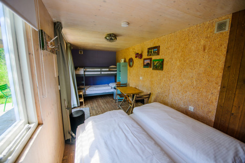 Pequeña casa luminosa con litera, cama doble, mesa comedor y grandes ventanas con vistas al exterior verde.