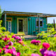 Pequeña casa de contenedor de envío con jardín florido y terraza de madera bajo el sol brillante.