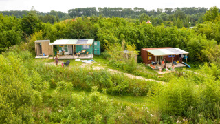 Casa pequeña de contenedor marítimo con fontanería, rodeada de naturaleza y personas en el exterior.