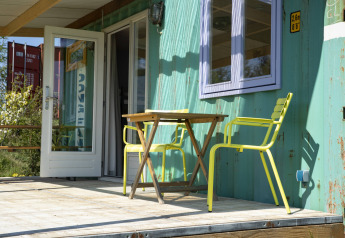 Solrig terrasse med gule stole og bord foran et shipping container tiny house på Netl Camping Kallumaan.