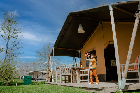 Bambina gioca con un giocattolo davanti a una tenda safari con servizi igienici, circondata dal verde.