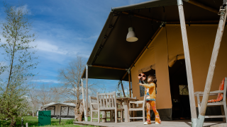 Kind speelt met speelgoed voor een safari tent met sanitair, omgeven door natuur en een zonnige hemel.