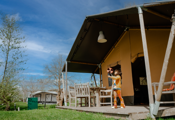 Enfant jouant avec un jouet devant une tente safari avec sanitaires, dans un cadre naturel et sous un ciel bleu.