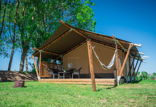 Tienda safari con terraza y hamaca en Camping de Tolbrug, Países Bajos, rodeada de árboles y cielo azul.