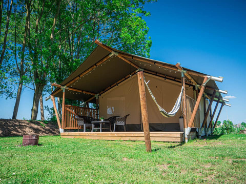 Safari-telt med terrasse og hængekøje ved Camping de Tolbrug, omgivet af grønne træer og blå himmel.