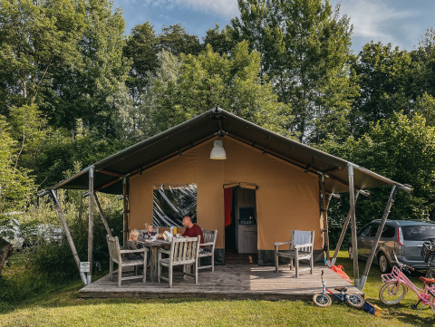 Familia sentada fuera de una tienda Safari con baño, rodeada de bicicletas y árboles en el camping.