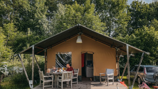 Gezin zit buiten bij een Safari tent met sanitair, omringd door fietsen en bomen op de camping.