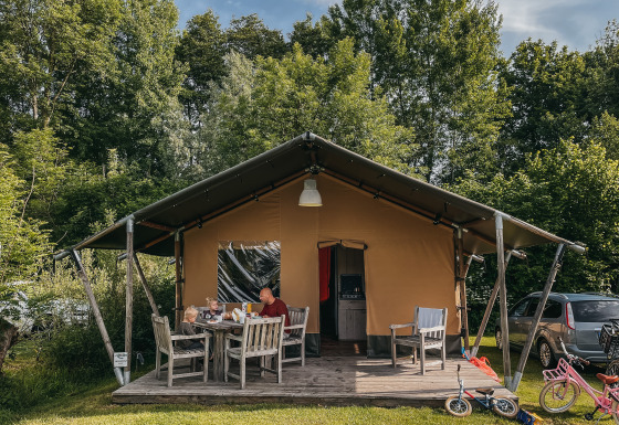 Famiglia seduta davanti a una tenda Safari con servizi igienici, circondata da bici e alberi in campeggio.