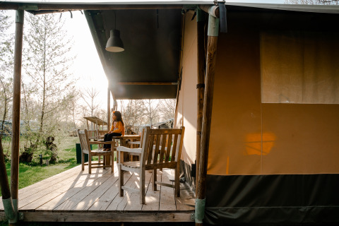 Frau sitzt auf Veranda eines Safarizelts mit Bad auf Campingplatz De Rammelbeek, Niederlande, bei Sonnenuntergang.