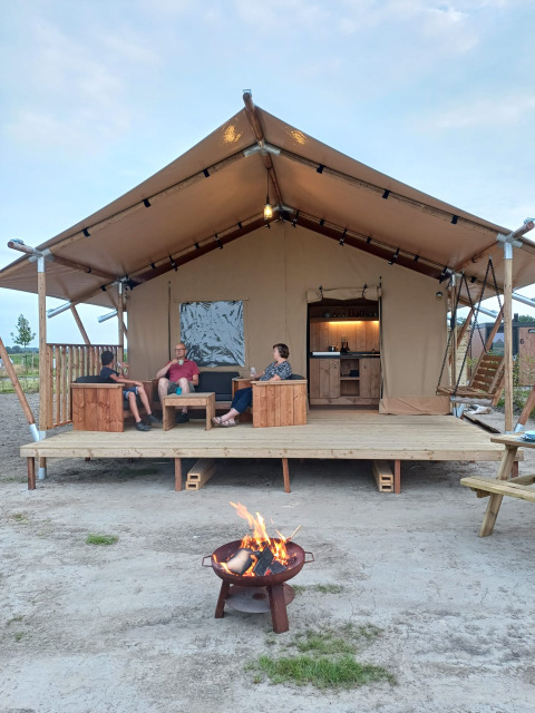 Mensen genieten op het terras van een safaritent met vuurkorf en comfortabele sanitaire voorzieningen.