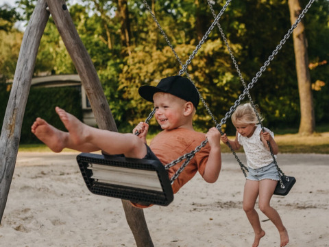 Zwei Kinder schaukeln lachend im Sand auf dem Spielplatz im Ferienpark Camping De Rammelbeek in Overijssel.