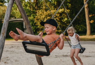 Zwei Kinder schaukeln lachend im Sand auf dem Spielplatz im Ferienpark Camping De Rammelbeek in Overijssel.