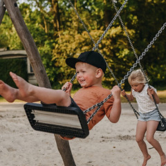 Zwei Kinder schaukeln lachend im Sand auf dem Spielplatz im Ferienpark Camping De Rammelbeek in Overijssel.