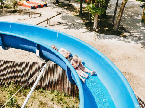 Dos personas bajan por un tobogán azul en un parque de vacaciones al aire libre rodeado de árboles y arena.
