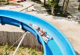 Zwei Personen rutschen eine blaue Wasserrutsche in einem Ferienpark mit sandigem Untergrund hinunter.