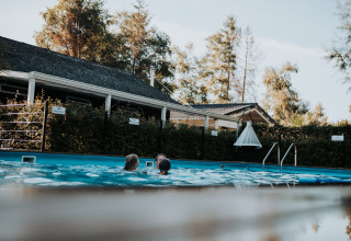 To personer svømmer i en udendørs pool ved Camping Emmen feriepark, Drenthe, Holland, omgivet af grønne træer.