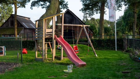 Playground with slide and swings at Camping Emmen, a holiday park in Drenthe, Netherlands, surrounded by trees.
