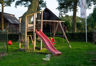 Playground with slide and swings at Camping Emmen, a holiday park in Drenthe, Netherlands, surrounded by trees.