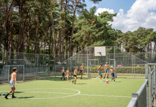 Bambini giocano a calcio su un campo esterno circondato da alberi alti a Familiepark Goolderheide, Belgio.