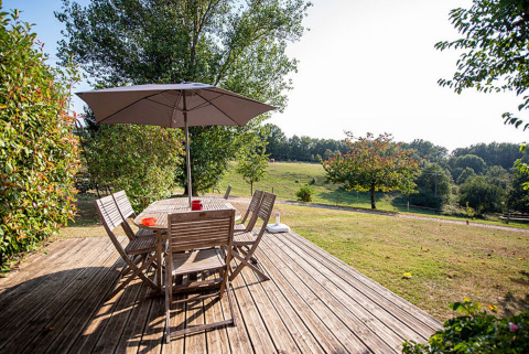 Wooden deck with table, chairs, and umbrella overlooking green fields at Camping Fargogne in Occitanie, France.