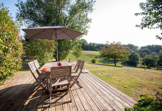 Wooden deck with table, chairs, and umbrella overlooking green fields at Camping Fargogne in Occitanie, France.