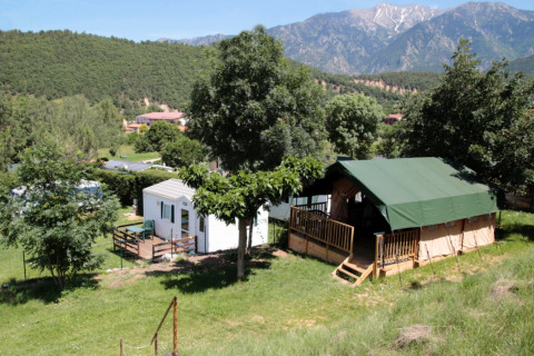 Zona de acampada en Camping le Rotja, Occitanie, Francia, con tienda y casa móvil rodeadas de montañas.