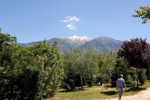 A man walks through the green Camping le Rotja park with mountain views and blue skies in Occitanie, France.