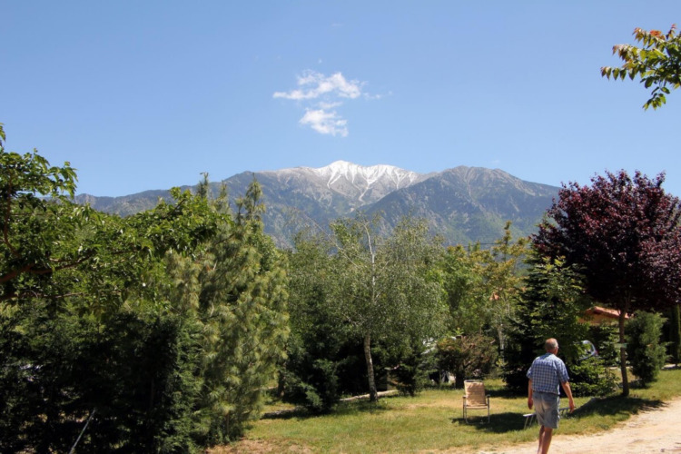 A man walks through the green Camping le Rotja park with mountain views and blue skies in Occitanie, France.