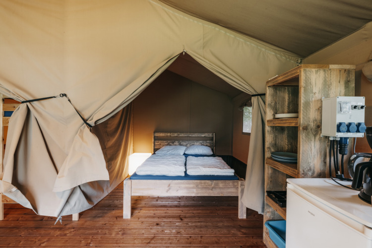 Innenaufnahme eines Safarizelts mit Holzbett, Küchenregal, Kühlschrank und Holzboden in Holland.