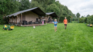 Two children play on a grassy lawn in front of a safari tent with toilet, surrounded by trees and toys.