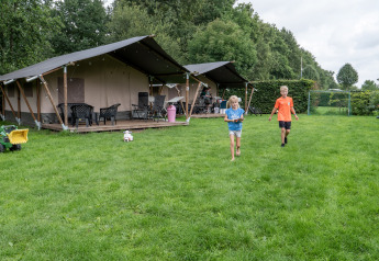 Due bambini giocano su un prato davanti a una tenda safari con bagno, circondati da alberi e giocattoli.