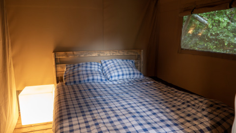 Slaapruimte in een safari tent met geruite lakens, houten bed en raamzicht op Camping Emmen in Nederland.