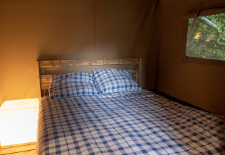 Indretning af en safari-telt med dobbeltseng, ternet sengetøj og vindue med udsigt til grønt ved Camping Emmen.