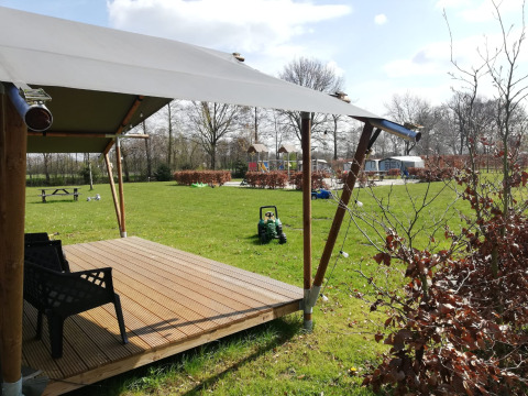 Tienda safari en Camping Emmen en los Países Bajos con terraza cubierta, césped y área de juegos al fondo.