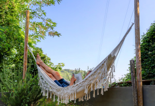 A person relaxes in a hammock and reads a book outdoors near the Safari tent at Camping Pian D'Amora in Italy.