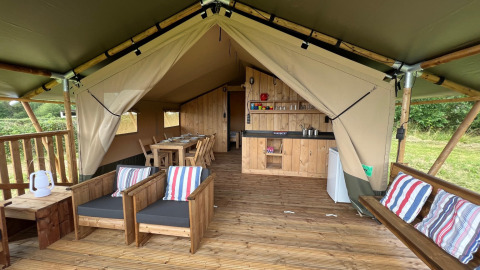 Interieur van een safaritent met houten zetels, eettafel en een compacte keukenhoek op het terras.